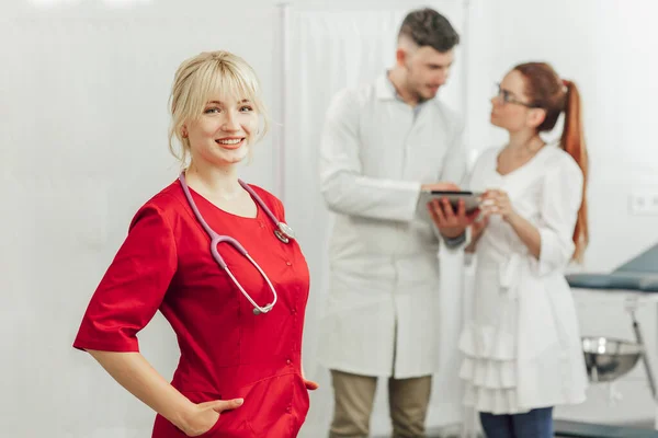 Közelkép egy mosolygó doktornőről, piros egyenruhában, sztetoszkóppal. Fiatal orvos mosolyog pózol a kamera előtt, mögötte a háttérben két másik orvos Stock Kép