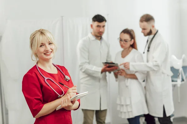 Dokter muda berseragam merah dengan buku catatan di kantor. Wanita melihat ke kamera. Di latar belakang, tim dari tiga dokter berbicara — Stok Foto