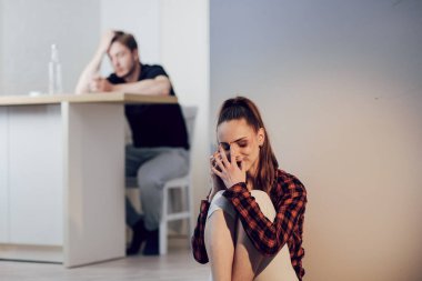 Aile içi şiddet kurbanı. Yüzünde morluklar olan korkmuş genç bir kadın mutfakta yerde oturan kız arkadaşına şikayet ediyor. Kocası arkada oturup içki içiyor.