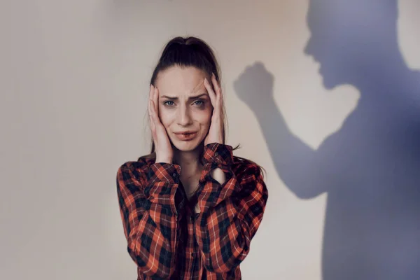 Violence against women. Portrait of young scared woman with a beaten face closes her ears with her hands. A large male shadow from the side shouts at the woman and threatens with a fist. Copy space — Stock Photo, Image