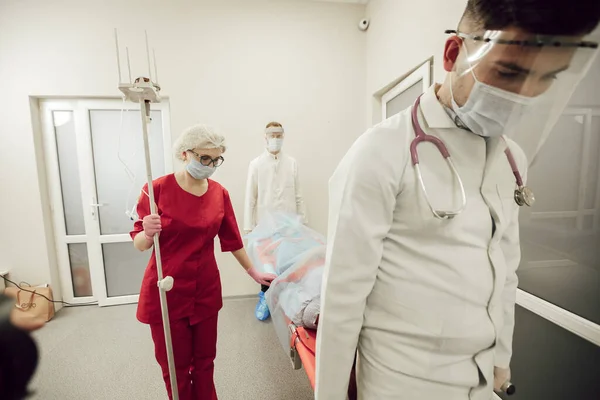 Un equipo de médicos lleva a un paciente enfermo en una camilla. Médicos de ambulancia con máscaras protectoras y trajes, el paciente sospecha de coronavirus. El trabajo de los médicos durante una pandemia — Foto de Stock