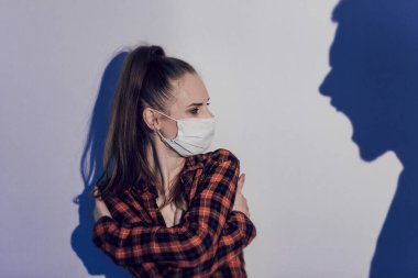Coronavirus karantinası sırasında kadınlara karşı şiddet. Gri arka planda izole edilmiş koruyucu maskeli yüzü dövülmüş, korkmuş genç bir kadın. Yandan büyük bir erkek gölgesi kadına bağırıyor.