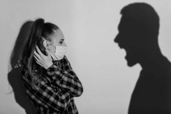 Violence against women during coronavirus quarantine. Young scared woman with a beaten face in a protective mask. Black and white. A large male shadow from the side shouts at the woman — Stock Photo, Image