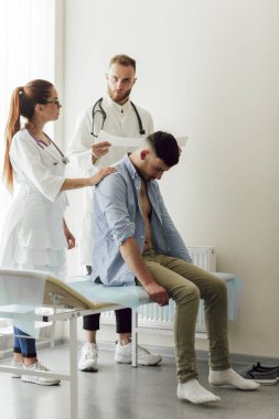 İki doktor, bir erkek ve bir kadın, hastane ofisinde genç bir hastayı muayene ediyorlar. Hastanede kanepede oturan genç bir adam.