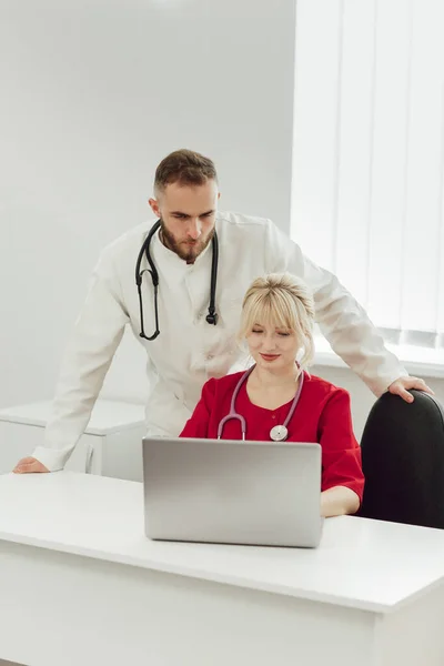 Dua dokter, seorang pria dan seorang wanita di kantor rumah sakit menggunakan laptop. Pekerja medis berseragam di rumah sakit berbicara satu sama lain — Stok Foto