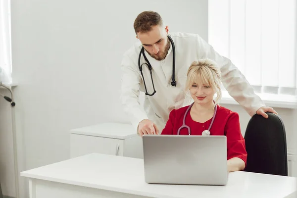 Dua dokter, seorang pria dan seorang wanita di kantor rumah sakit menggunakan laptop. Pekerja medis berseragam di rumah sakit berbicara satu sama lain — Stok Foto
