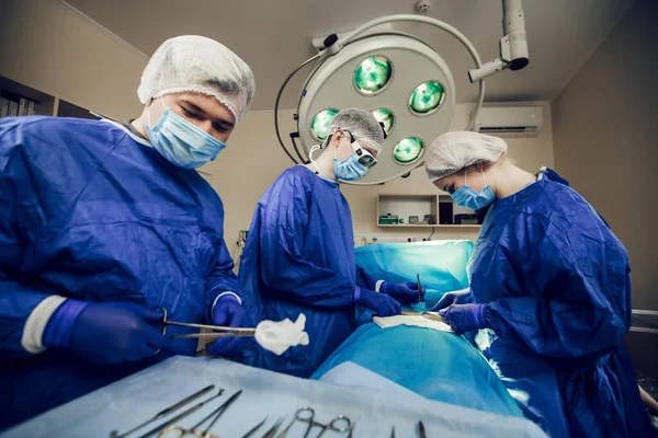 Een team chirurgen in een blauw uniform opereert een patiënt in een ziekenhuis. Zijaanzicht van jonge artsen op het werk. Een jonge arts desinfecteert chirurgische schaar — Stockfoto