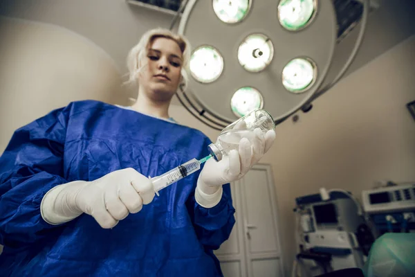 Jonge blonde arts in witte handschoenen met spuit en medicijnfles in de operatiekamer. Medicijnconcept. De arts neemt het geneesmiddel in een spuit. Voorbereiding voor chirurgie, verdoving — Stockfoto