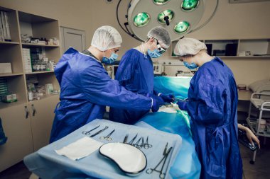 Mavi üniformalı bir cerrah ekibi hastanede bir hastayı ameliyat ediyor. İşteki genç doktorların yan görünüşü