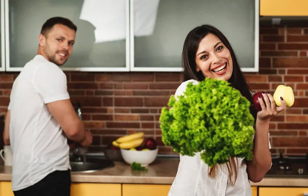 Pasangan muda memasak di dapur. Wanita tersenyum, memegang selada dan apel. Pria di latar belakang. Makan Sehat — Stok Foto