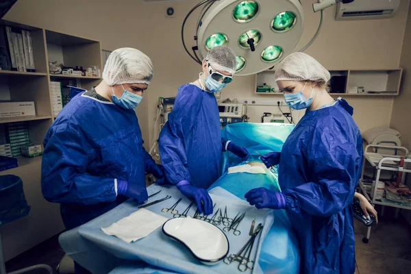 Sebuah tim ahli bedah dengan seragam biru beroperasi pada pasien di rumah sakit. Pandangan samping dokter muda di tempat kerja. Seorang dokter muda mensterilkan gunting bedah — Stok Foto