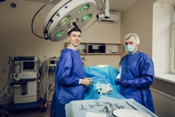 Dua dokter muda berseragam di ruang operasi. Dua ahli bedah dan seorang pasien di rumah sakit sedang mempersiapkan operasi, para pria sedang melihat ke kamera — Stok Foto