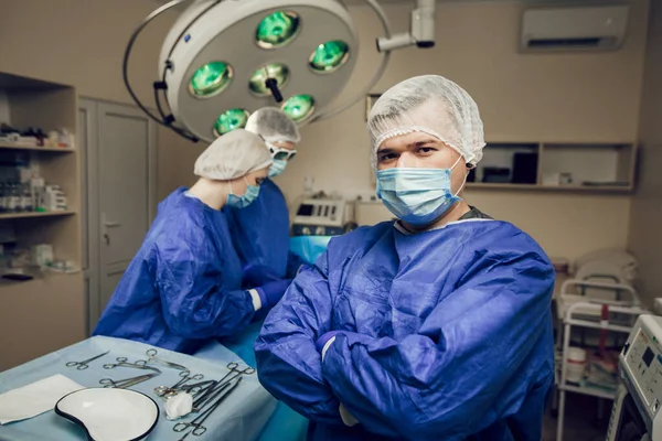 Potret seorang ahli bedah dalam topeng pelindung di ruang operasi. Di belakangnya, dua dokter lain sedang mempersiapkan operasi — Stok Foto