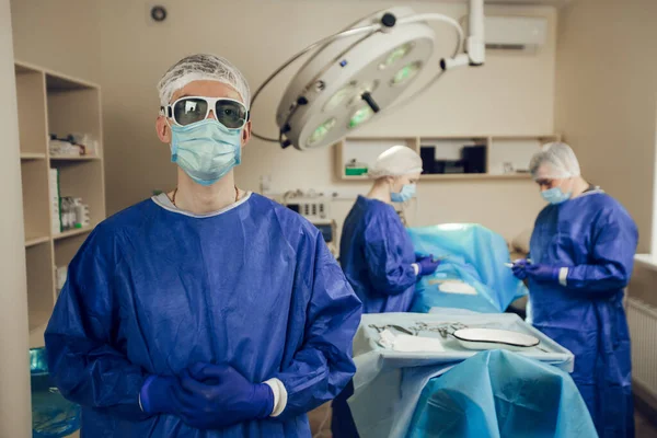 Potret seorang ahli bedah dengan topeng pelindung dan kacamata di ruang operasi. Di belakangnya, dua dokter lain sedang mempersiapkan operasi — Stok Foto