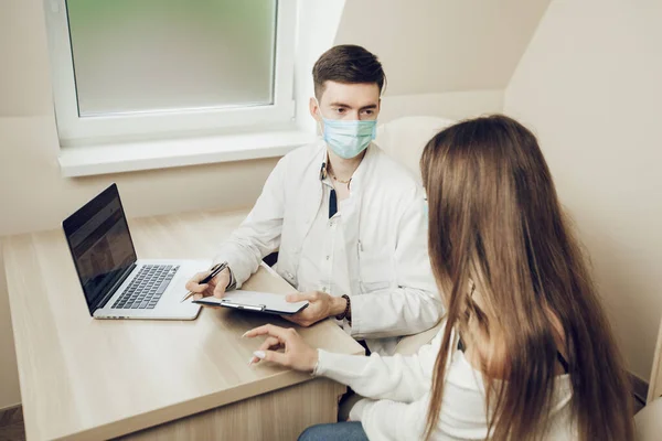Masuk ke dokter selama karantina. Dokter muda dan pasien duduk di kantor mengenakan masker pelindung. — Stok Foto