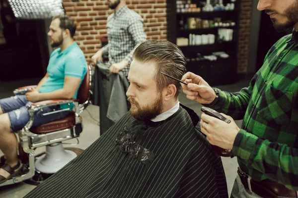 Corte de cabelo na barbearia. Vista lateral do jovem barbudo recebendo corte de cabelo enquanto sentado na cadeira na barbearia. Cabeleireiro hidrata o cabelo com spray — Fotografia de Stock