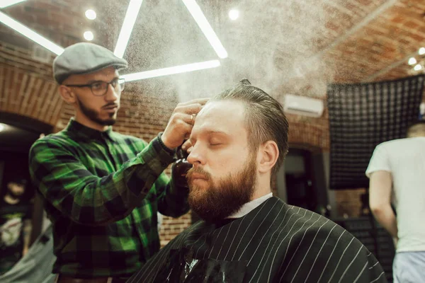 Corte de cabelo na barbearia. Vista lateral do jovem barbudo recebendo corte de cabelo enquanto sentado na cadeira na barbearia. Cabeleireiro hidrata o cabelo com spray — Fotografia de Stock