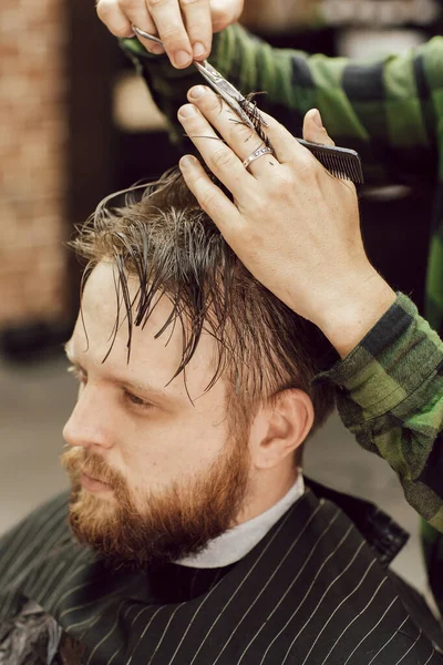 Corte de cabelo na barbearia de perto. O cabeleireiro corta o jovem barbudo com um pente e uma tesoura — Fotografia de Stock