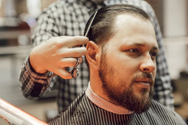 Corte de cabelo na barbearia de perto. O cabeleireiro corta o jovem barbudo com um pente e uma tesoura — Fotografia de Stock