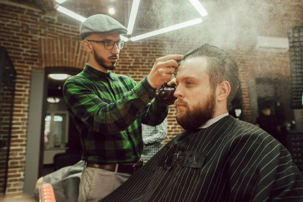 Corte de cabelo na barbearia. Vista lateral do jovem barbudo recebendo corte de cabelo enquanto sentado na cadeira na barbearia. Cabeleireiro hidrata o cabelo com spray — Fotografia de Stock