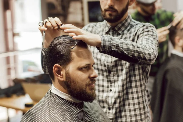 Corte de cabelo na barbearia. Vista de perto do jovem barbudo cortando o cabelo enquanto estava sentado na cadeira da barbearia. Cabeleireiro corta cabelo com tesoura e um pente — Fotografia de Stock