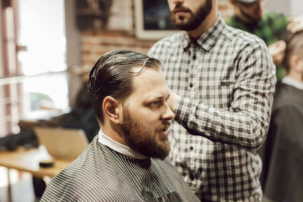 理发店理发。坐在理发店的椅子上，近距离观察年轻的留着胡子的男人理发的情景。理发师用剪刀和梳子剪头发 — 图库照片