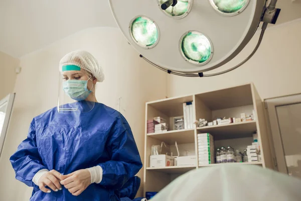 Seorang dokter bedah wanita muda dalam topeng pelindung, kacamata dan perisai di ruang operasi. Seorang dokter di rumah sakit selama pandemi coronavirus. Operasi selama karantina — Stok Foto