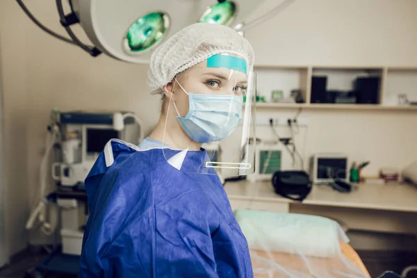 Potret seorang dokter bedah wanita muda dengan topeng pelindung, kacamata dan perisai di ruang operasi. Seorang dokter di rumah sakit selama pandemi coronavirus. Operasi selama karantina — Stok Foto