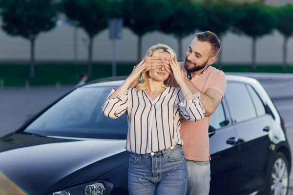 남자는 새 차를 구입 함으로써 여자를 놀라게 한다. 젊은 부부가 자동차를 삽니다. 한 남자와 여자가 길에서 차 근처에 서 있습니다. 사랑하는 사람에게는 뜻밖의 선물 — 스톡 사진