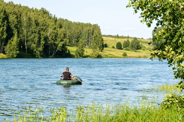 A man swims in an inflatable boat on the river against the background of forests and meadows
