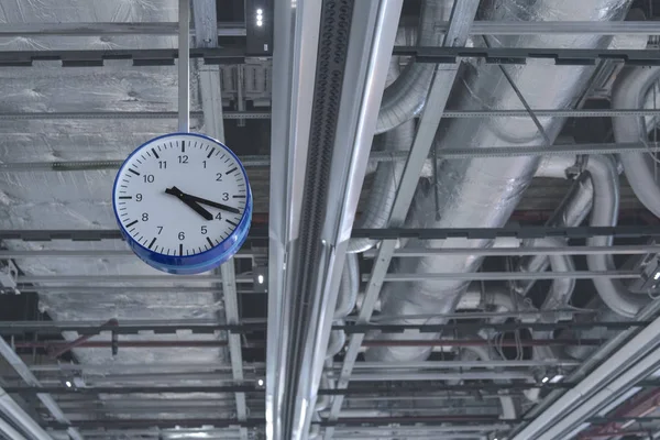 View of the clock that hangs from the ceiling against the background of ventilation pipes. — Stock Photo, Image