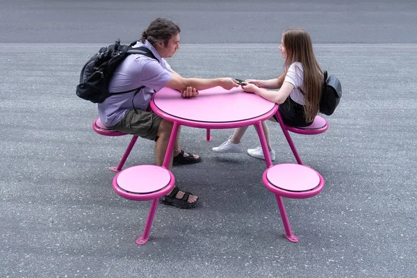 A young man and girl are sitting at a pink table in the street.