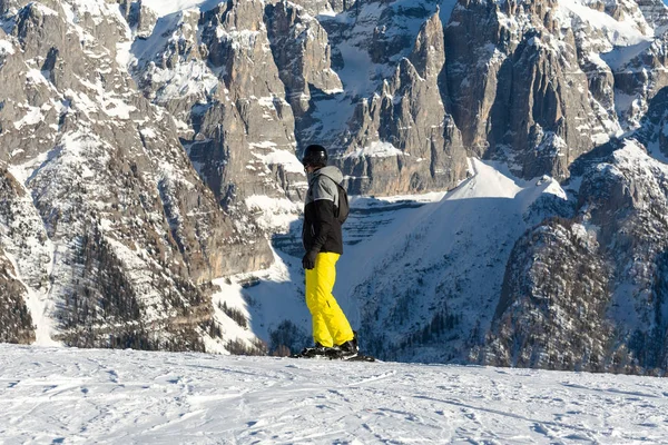 Um snowboarder em uma jaqueta preta e calças amarelas está em uma inclinação de esqui contra o pano de fundo da montanha . — Fotografia de Stock