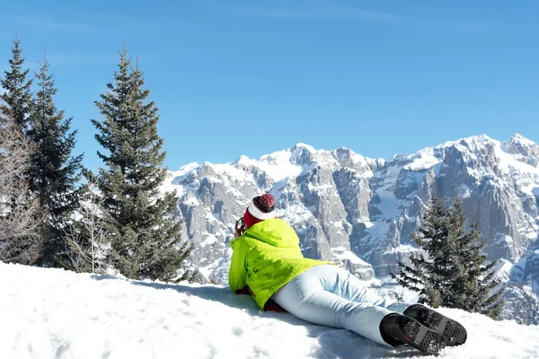 Yeşil ceketli kız snowboardcu bir yamaç üzerinde yatıyor ve dağlara bakıyor. 