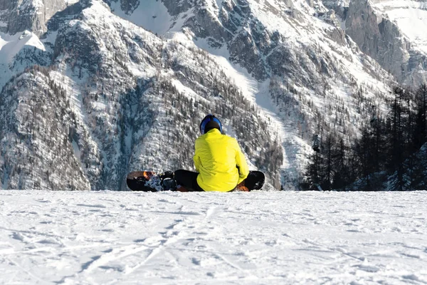 Snowboardista ve žlutém saku sedí na lyžařském svahu na pozadí hory. — Stock fotografie