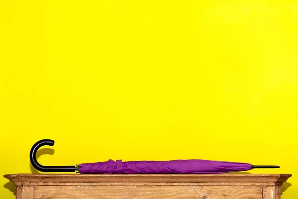 A folded umbrella in purple lies on a wooden chest of drawers against a yellow wall. Concept interior, order in the house.