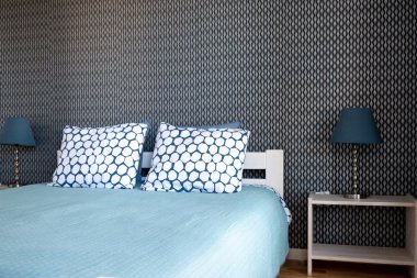 Koyu duvara ve lambalı komilere karşı mavi yatak örtüsü ve beyaz yastıklı çift kişilik yatak görünümü. 