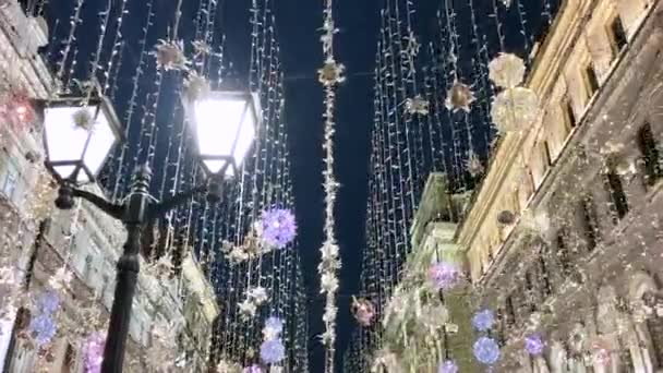 La calle está decorada con guirnaldas luminosas de luces que se balancean en el viento sobre el fondo del cielo oscuro y los edificios. Moscú. Calle Nikitskaya. — Vídeo de stock