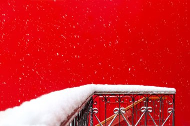 Kar yağışı sırasında binanın kırmızı duvarına karşı dövülmüş metalden yapılmış karlı balkon ve teras manzarası. Mimarlık, kış, kartpostal, Noel, ilgi