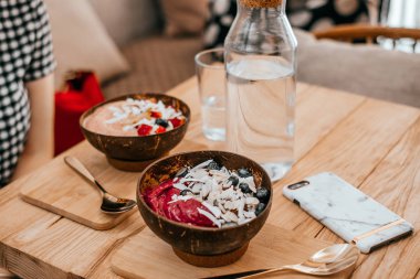 Kiev, Ukrayna - 19 Ağustos 2018: İki güzel kase pembe berry smoothie muz, çilek, ahududu, yaban mersini, Hindistan cevizi ve badem smartphone mermer durumda ahşap tablo ile.