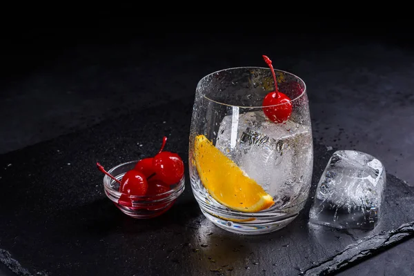 Ingredients for Cocktail on a black slate background. Gass with ice cube, orange slice and cherry with copy space on a stone background.