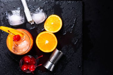 Tekila Sunrise kokteyli, portakal, buz küpleri, maraschino kirazı, buz maşaları ve aletlerden bir ıslak siyah arduvaz tepsi. Kopya alanı ile üstten görünüm.