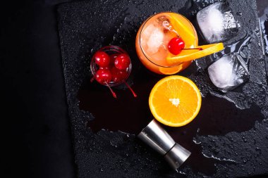 Tekila Sunrise kokteyli, portakal, buz küpleri, maraschino kirazı ve aletlerden bir ıslak siyah arduvaz tepsi. Kokteyl küme. Üstten görünüm. Metin alanı olan üstten görünüm.