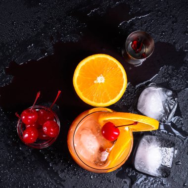 Tekila Sunrise kokteyli, portakal, buz küpleri, maraschino kirazı ve aletlerden bir ıslak siyah arduvaz tepsi. Üstten görünüm.