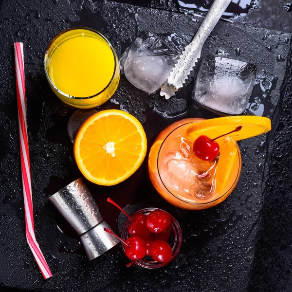 Tequila Sunrise cocktail, orange, ice cubes, maraschino cherries, ice tongs, jigger and straw on a black wet slate tray. Top view.