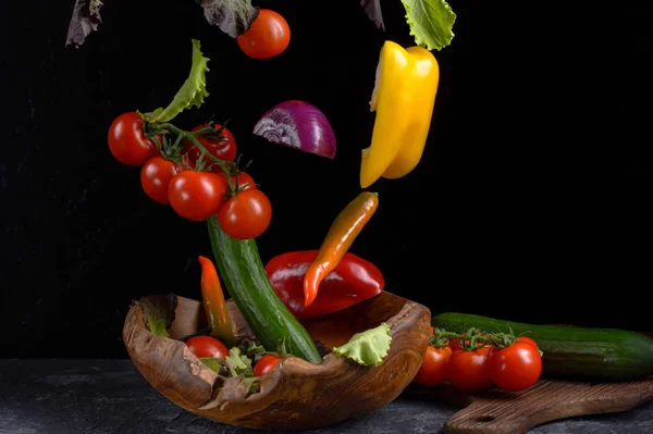 Salatzutaten schweben. gesunde vegetarische Kost fliegt über eine hölzerne Schüssel auf einem dunklen Steintisch mit Schneidebrett und dunklem Hintergrund.. — Stockfoto