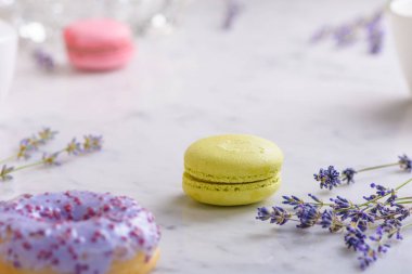 Görüntünün ortasında bir antep fıstığı makarna ve sprigs lavanta, önünde bir leylak donut, arkasında bir pembe makarna, beyaz mermer masa üzerinde