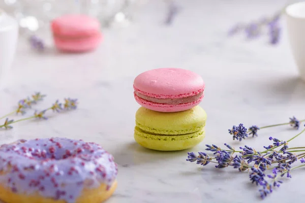 Two macarons and sprigs lavender in the center of the image, one lilac donut in front, one pink macaron behind, on white marble table