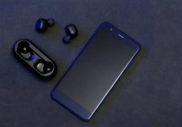 Black wireless headphones with blue cellphone on dark background. Flat lay view of modern wireless earphones with charging case. TWS headphones near charging case.