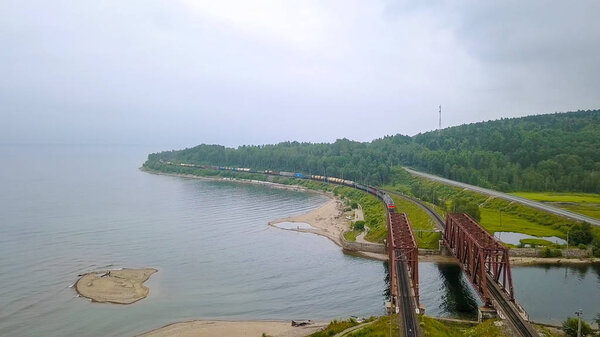 Транссибирская железная дорога, побережье Байкала. Движение поездов по железному мосту через реку, впадающую в Байкал. Россия, с дрона
  